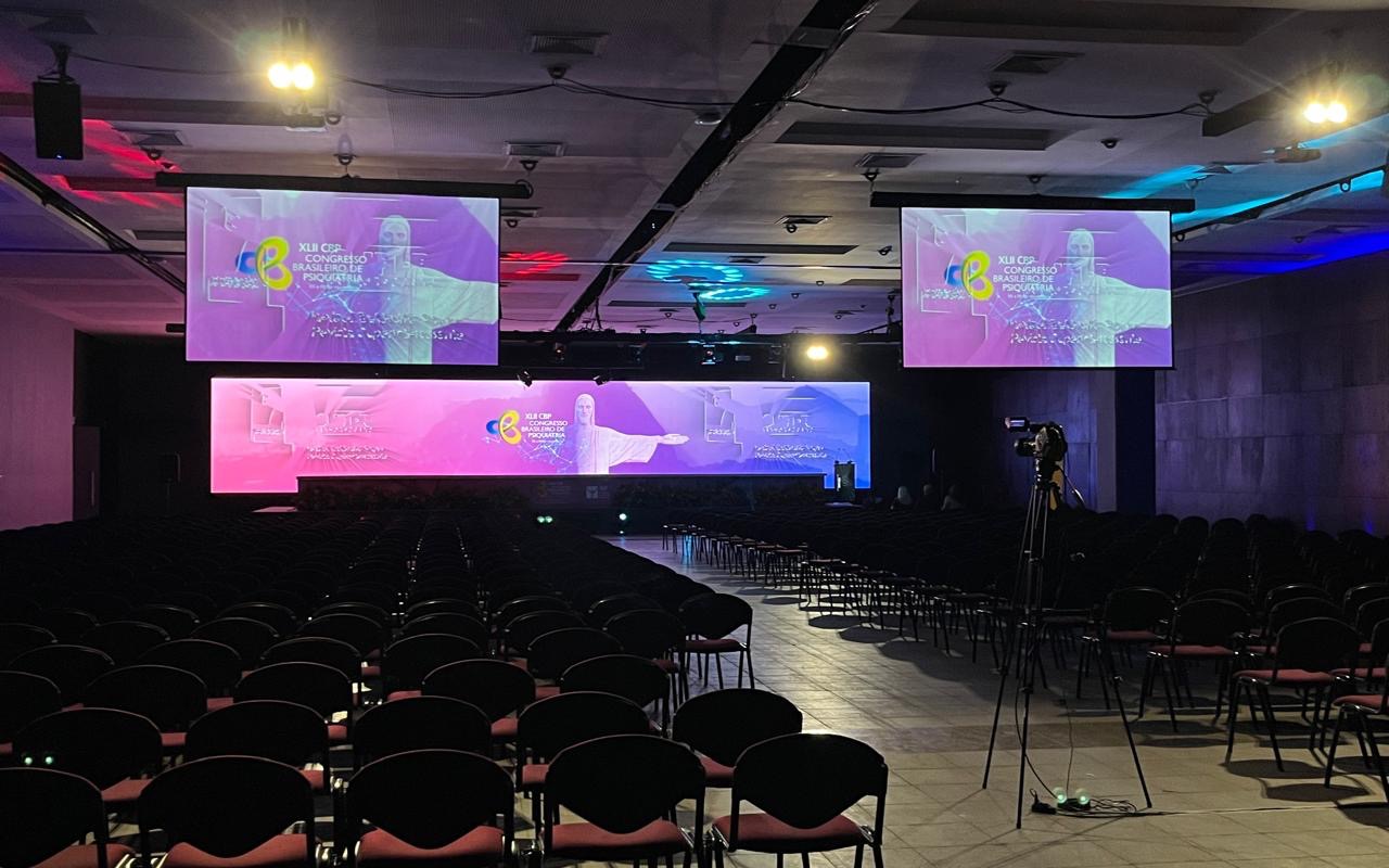 Auditório de congresso vazio com duas telas de projeção acesas, painel de LED cobrindo a frente do palco e câmera em tripé ao fundo