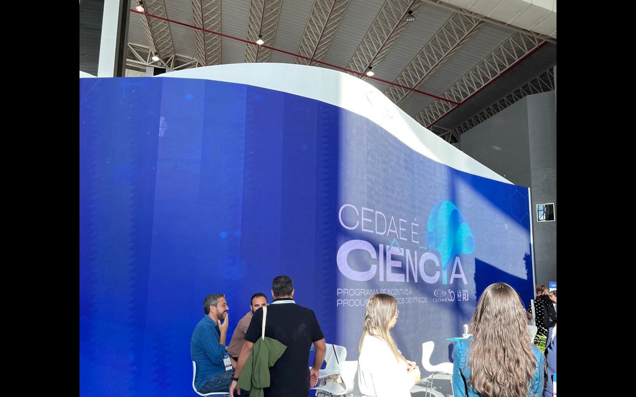 Parede curva de LED azul recortada em onda ocupa a frente do estande, com lounge de banquetas na base e treliça do pavilhão acima