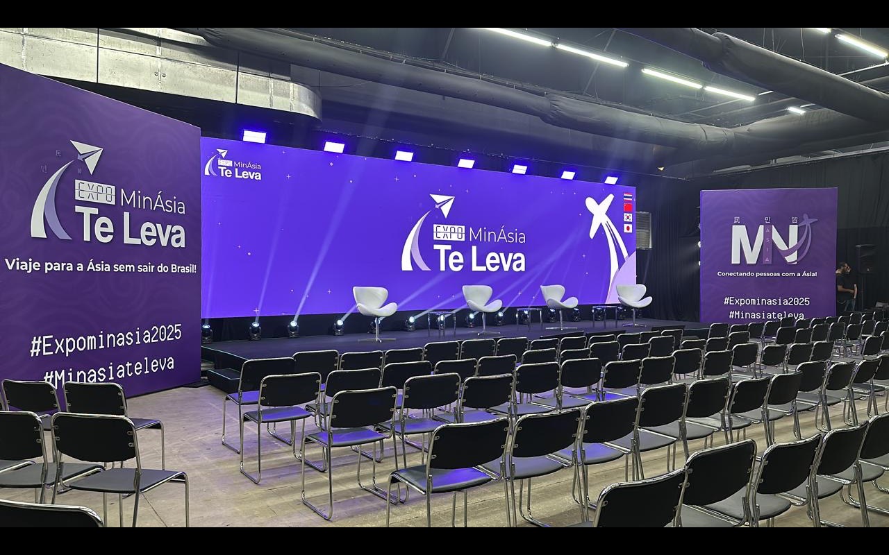 Fileiras de cadeiras vazias voltadas para o palco com quatro poltronas brancas, painel de LED roxo ao fundo e refletores no piso do palco