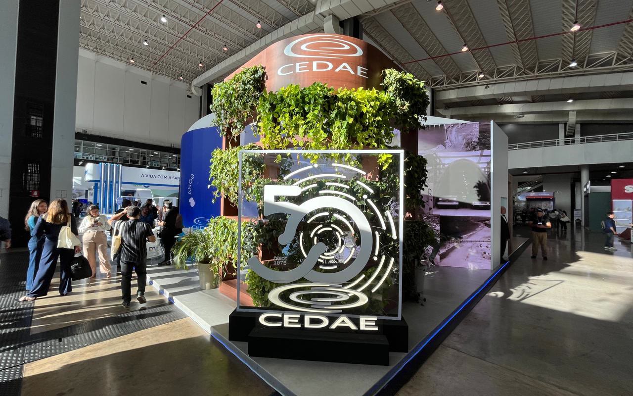 Estande em cubo com logo iluminado da CEDAE, jardim vertical e painel gráfico lateral, visitantes circulando sob treliça do pavilhão