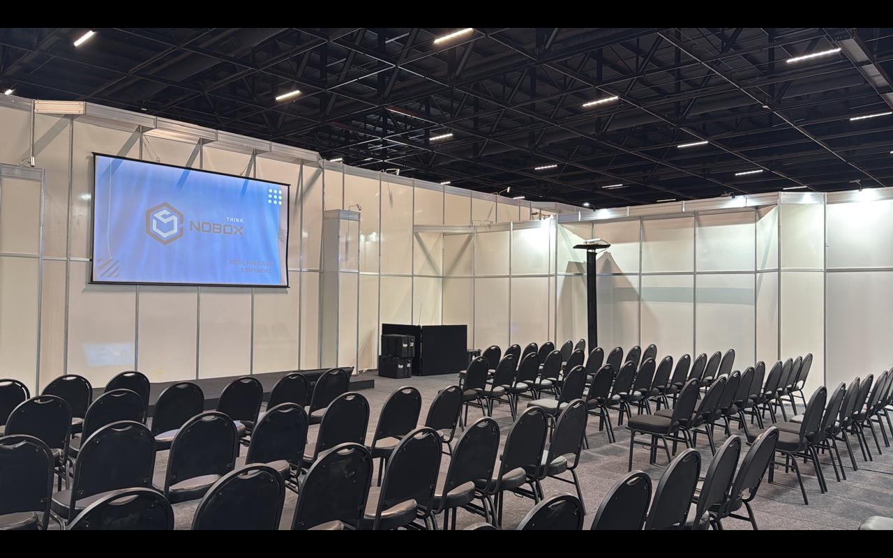 Sala de palestras montada dentro de pavilhão, cadeiras vazias em fileiras, tela de projeção acesa e estrutura metálica do teto aparente