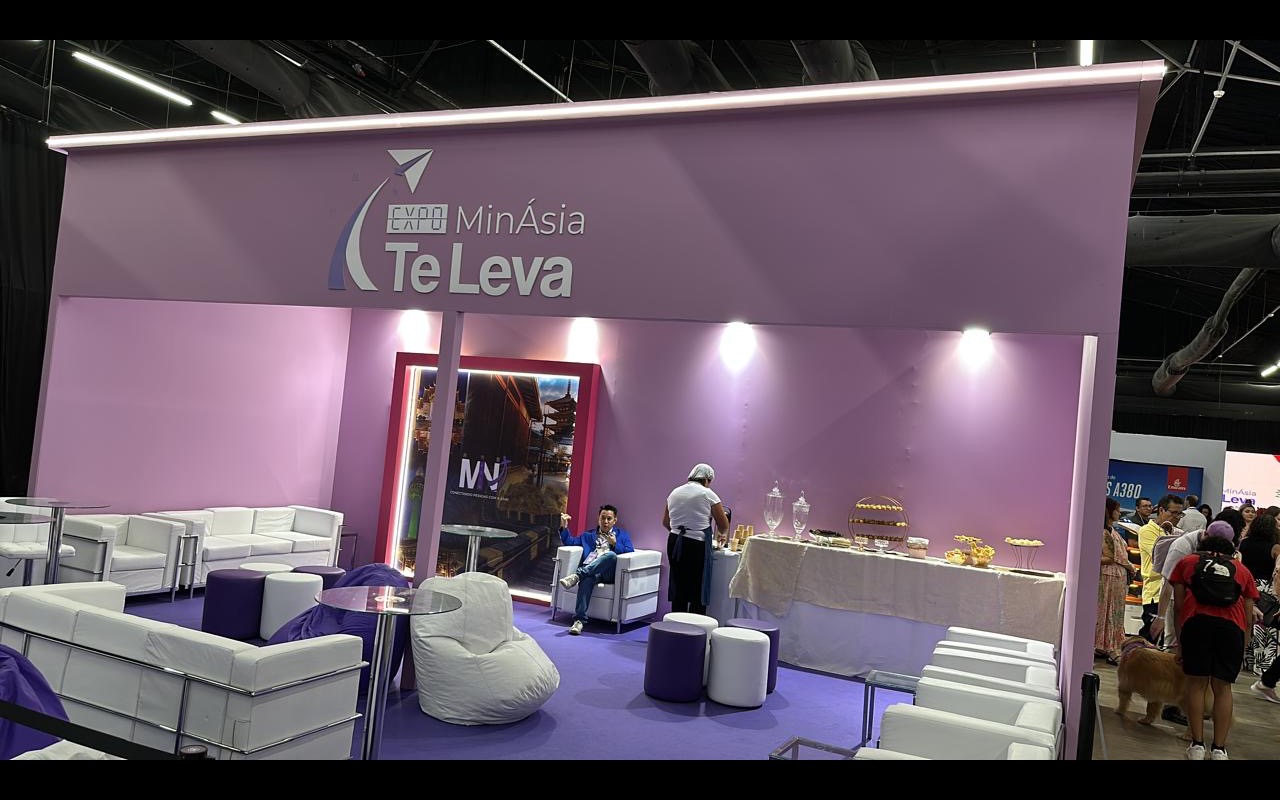 Estande com testeira iluminada e spots acesos sobre lounge de sofás brancos, carpete roxo, mesa de buffet e teto técnico do pavilhão.