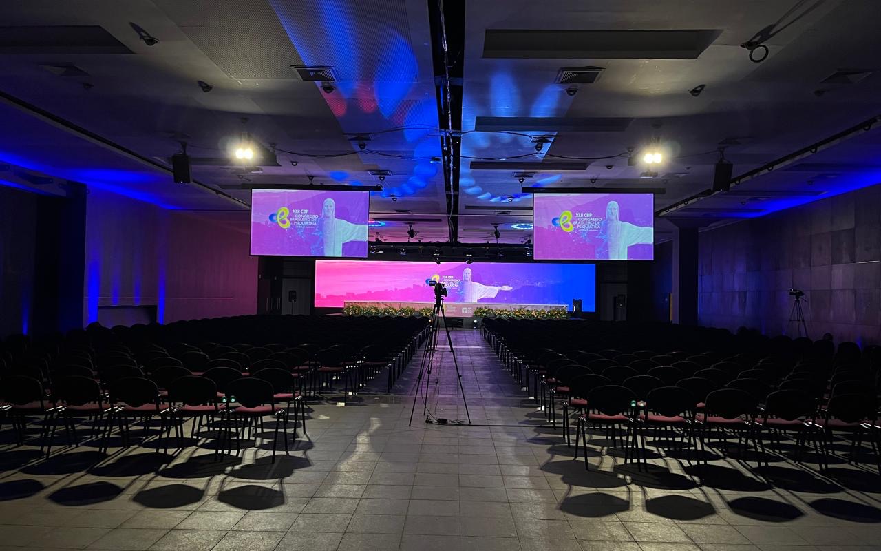 Fileiras de cadeiras vazias diante de palco com painel de LED, duas telas suspensas em treliça e câmera montada em tripé no corredor central