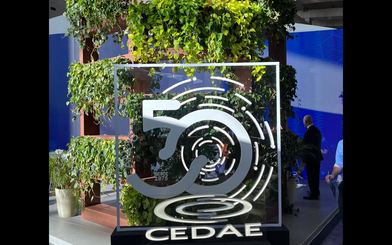 Parede de plantas em prateleiras de madeira e escultura vazada dos 50 anos da CEDAE sobre base preta ocupam a frente do estande.