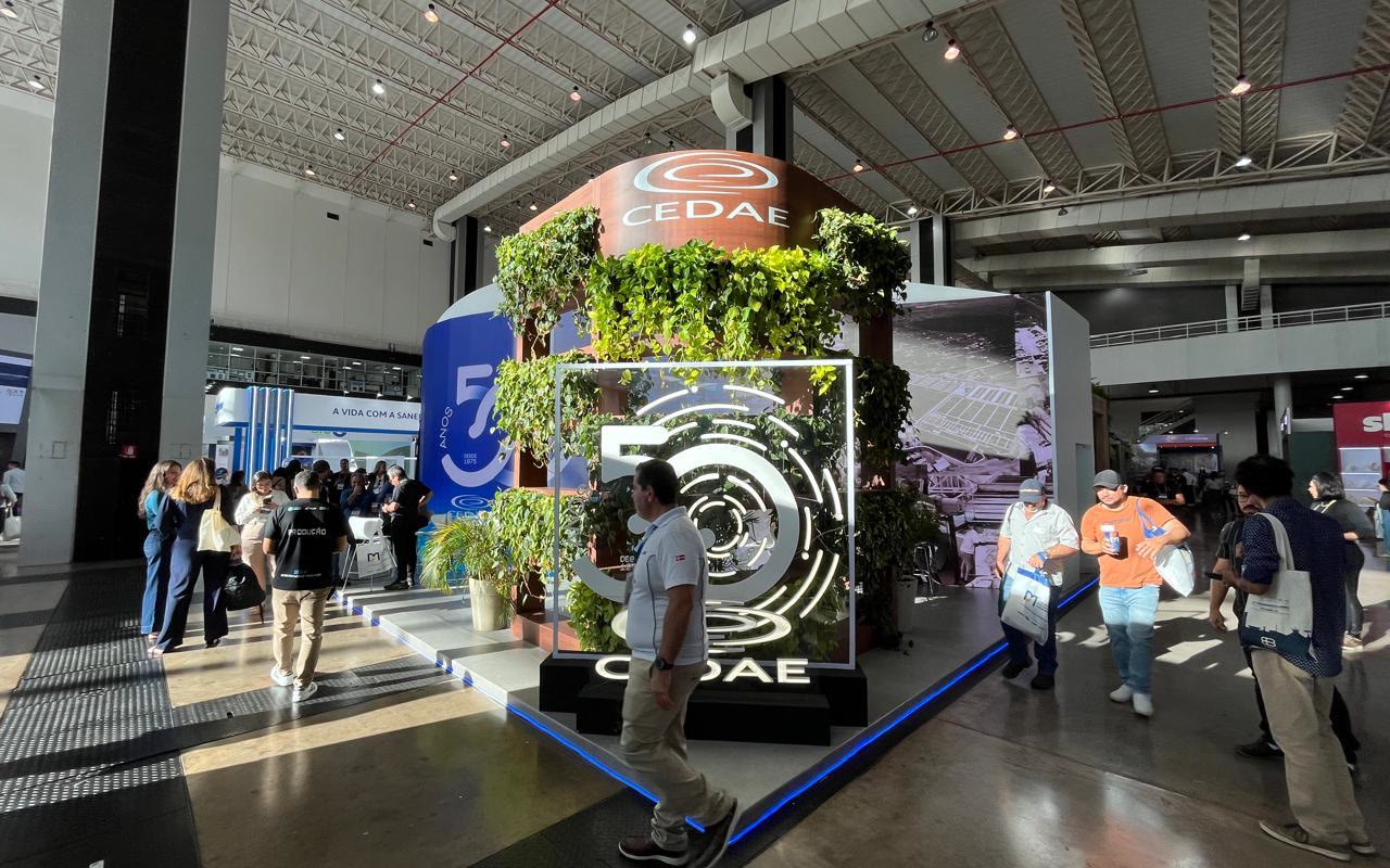 Torre curva revestida de plantas no centro do estande, cubo de luz com o número 50, piso elevado com fita de LED azul e visitantes passando