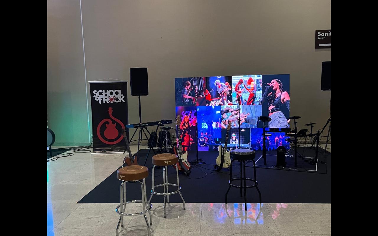 Cenário de gravação montado na área de circulação do evento: três banquetas voltadas para um painel de LED, guitarras em pedestais e caixa de som.