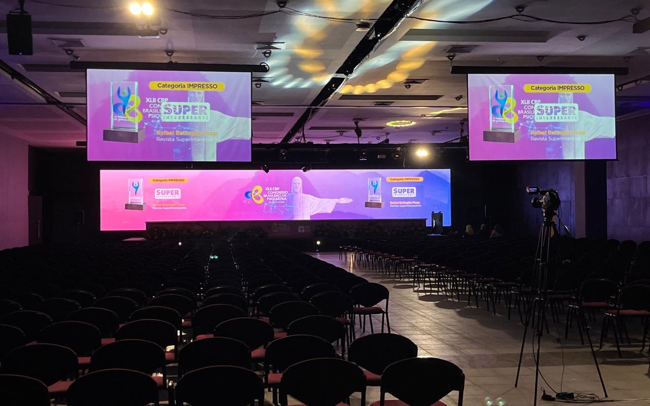 Auditório de congresso com fileiras de cadeiras vazias, dois telões suspensos, painel de LED no palco e câmera em tripé à direita