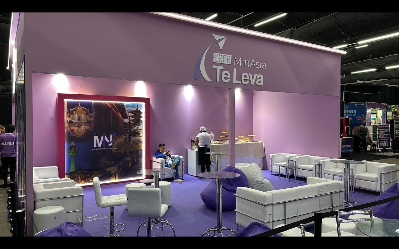 Estande de feira com parede lilás lavada por spots, friso de luz no topo, painel retroiluminado e lounge de poltronas brancas em carpete roxo