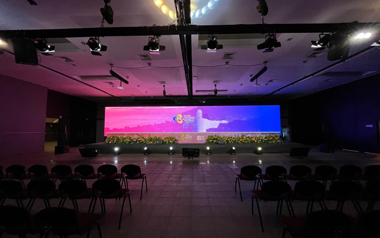 Palco de congresso com parede de LED acesa, jardineiras floridas na boca de cena, treliça de refletores no teto e cadeiras vazias em fileiras