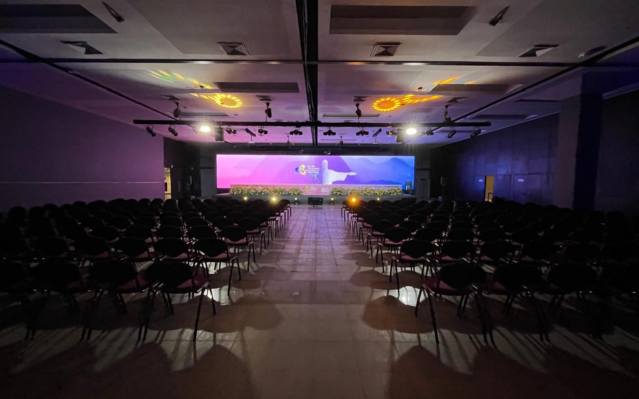 Plenária vazia já montada, fileiras de cadeiras até o fundo, painel de LED aceso ao centro do palco e treliça com refletores no teto