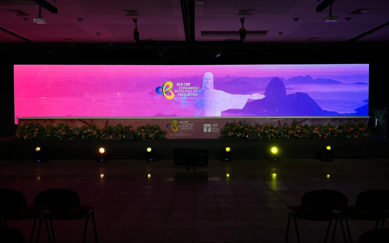 Painel de LED panorâmico cobrindo toda a largura do palco, arranjo de flores na borda, fila de refletores no piso e cadeiras vazias em primeiro plano