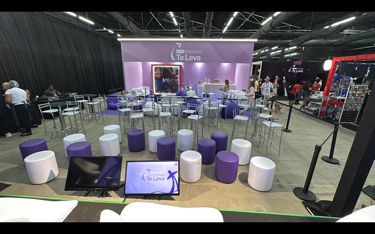 Área de lounge de feira com banquetas altas, pufes roxos e brancos, tela acesa emoldurada no fundo cenográfico e dois monitores no piso