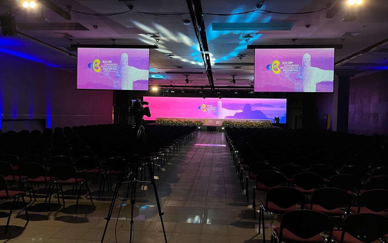 Plenária montada e vazia, fileiras de cadeiras ao longo do corredor central, painel de LED no palco, telas suspensas e câmera em tripé no fundo da sala