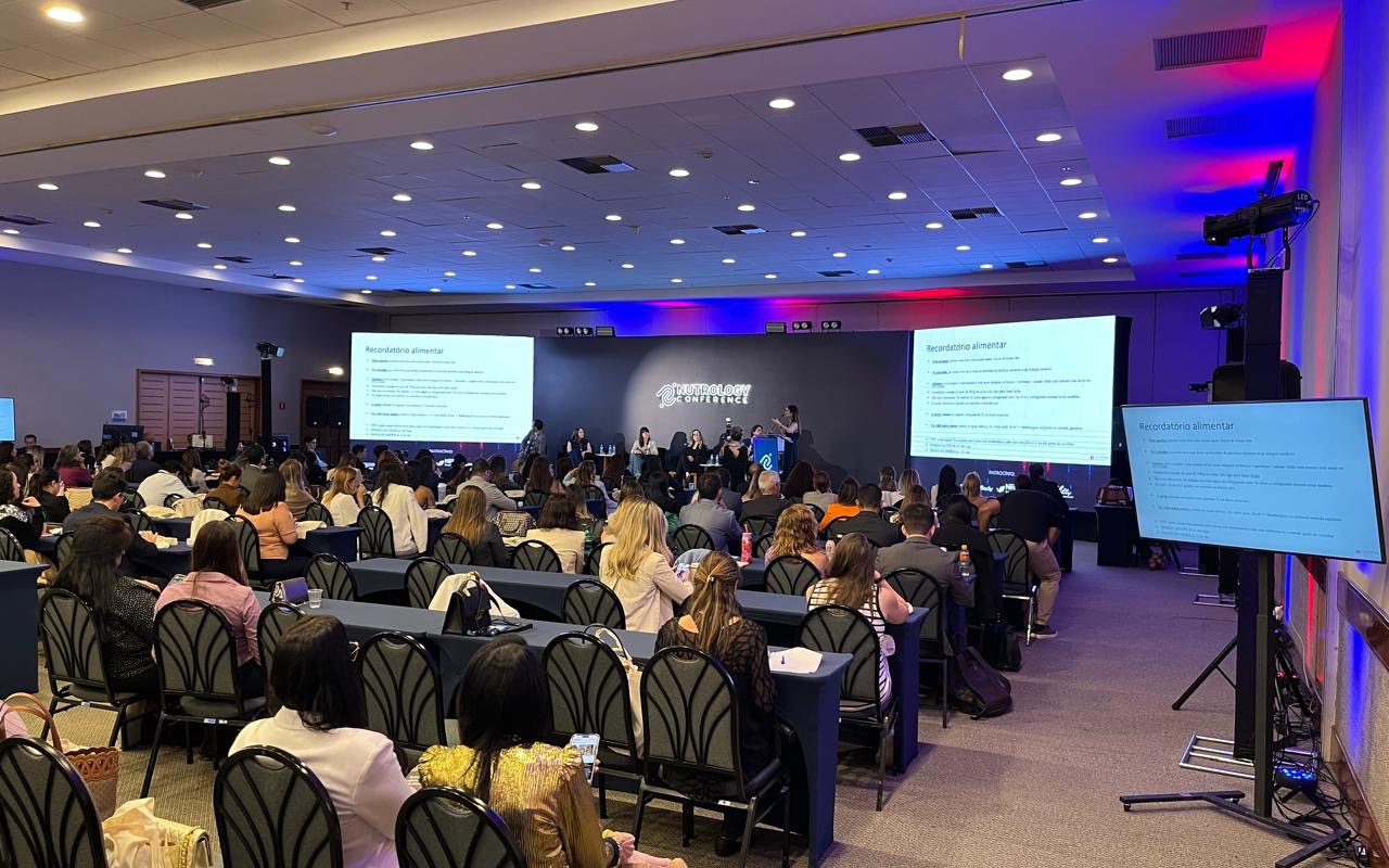 Plateia sentada em mesas longas de frente para o palco, duas telas de projeção laterais, mesa de debate no palco e monitor de retorno em primeiro plano