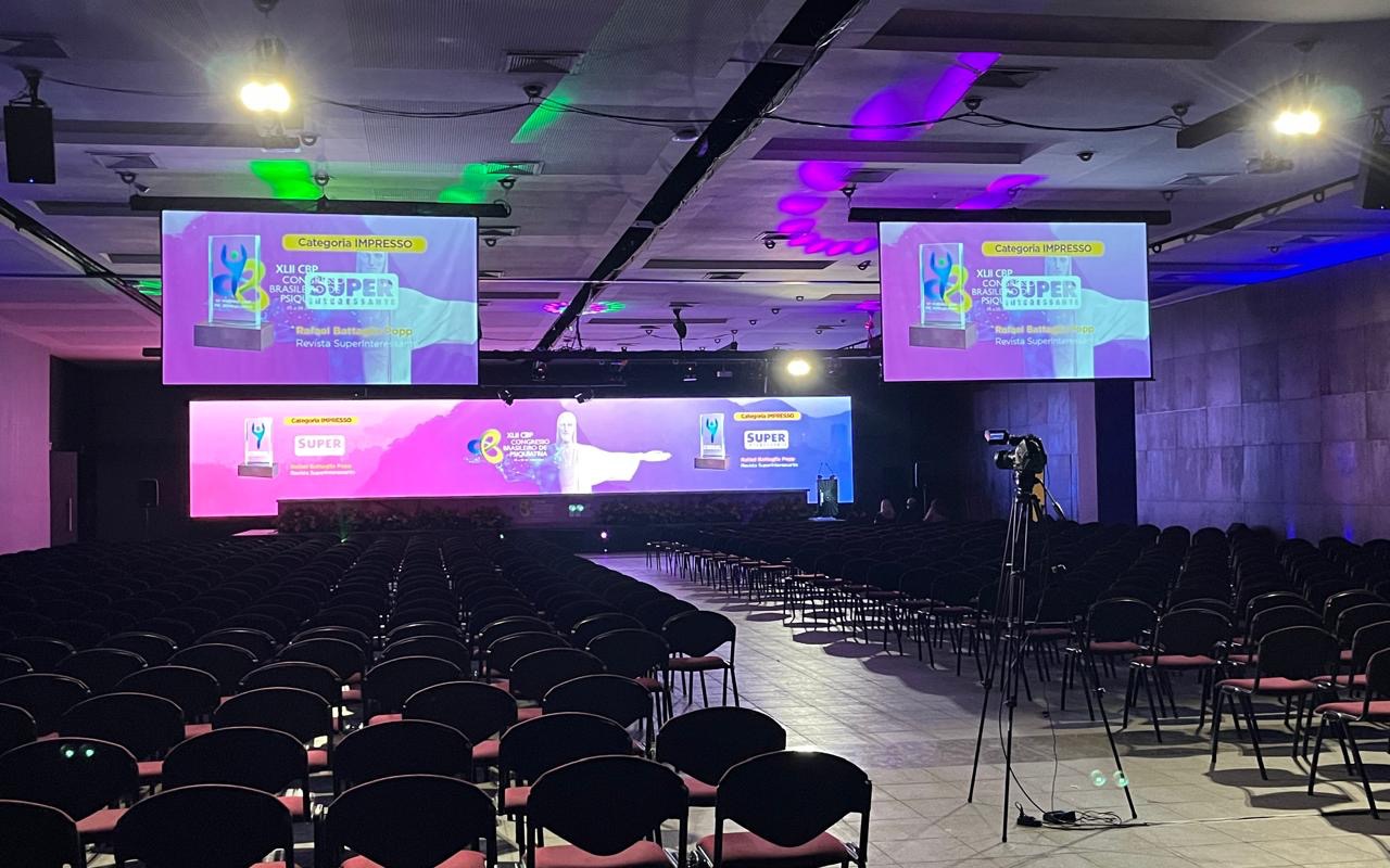 Auditório de congresso com fileiras de cadeiras vazias diante do palco com parede de LED, dois telões suspensos e câmera em tripé com cabo no piso
