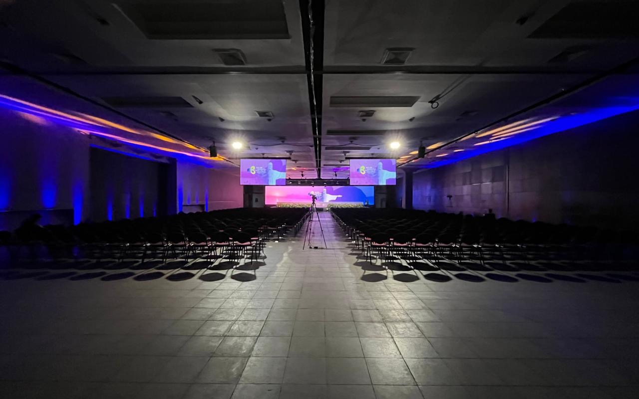 Plenária de congresso com as cadeiras ainda vazias, duas telas suspensas, painel de LED no palco e câmera em tripé no corredor central