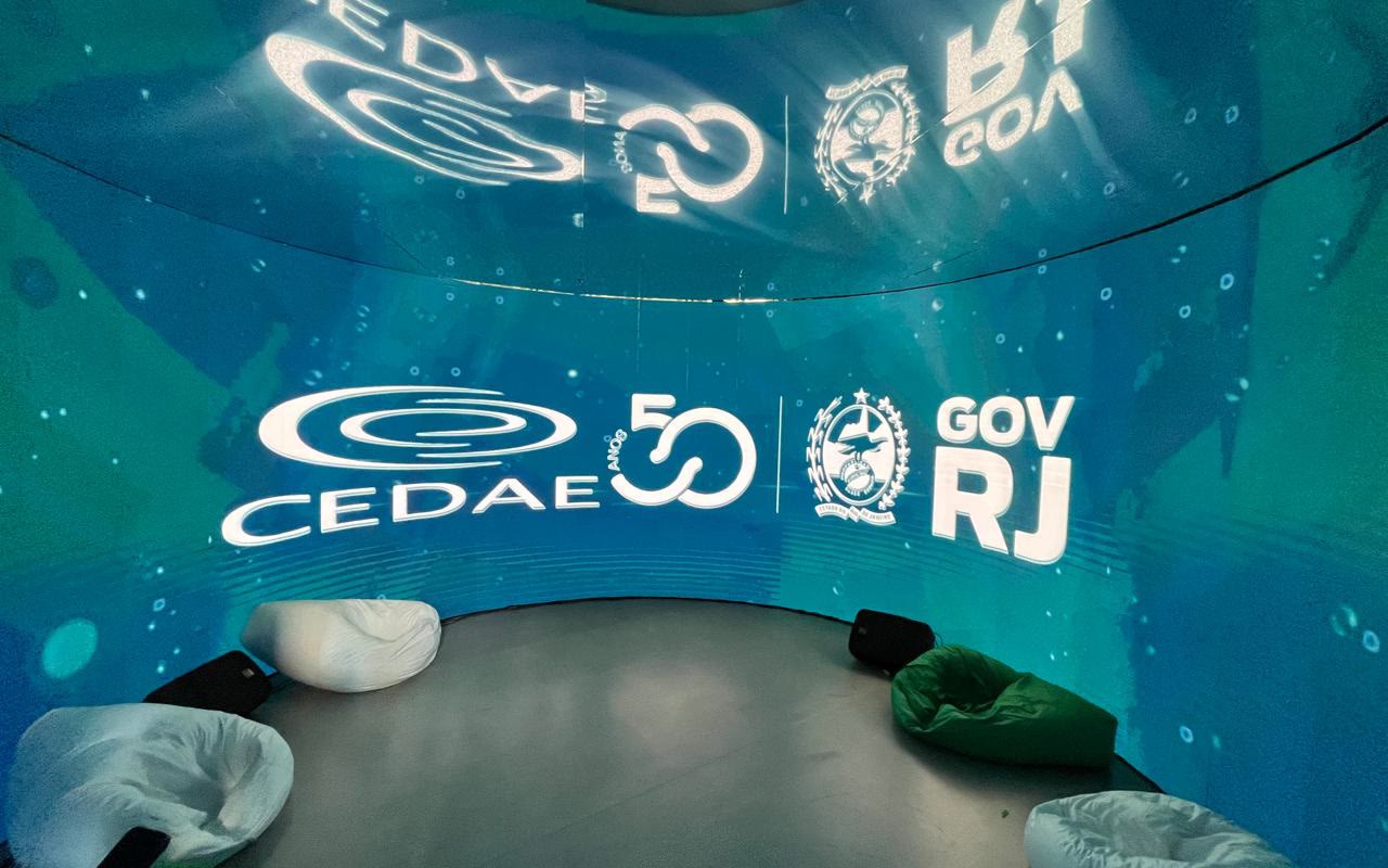 Parede curva de LED em azul e verde dentro de estande, marca CEDAE ao centro, pufes brancos e verdes e caixas de som rente ao piso