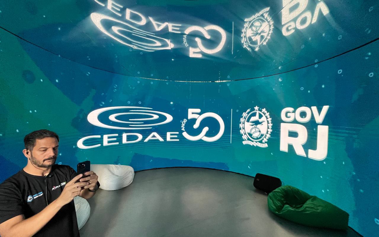 Parede de LED curva com a marca CEDAE 50 anos e Governo do RJ, teto espelhado refletindo o painel e um operador de pé com o celular na mão
