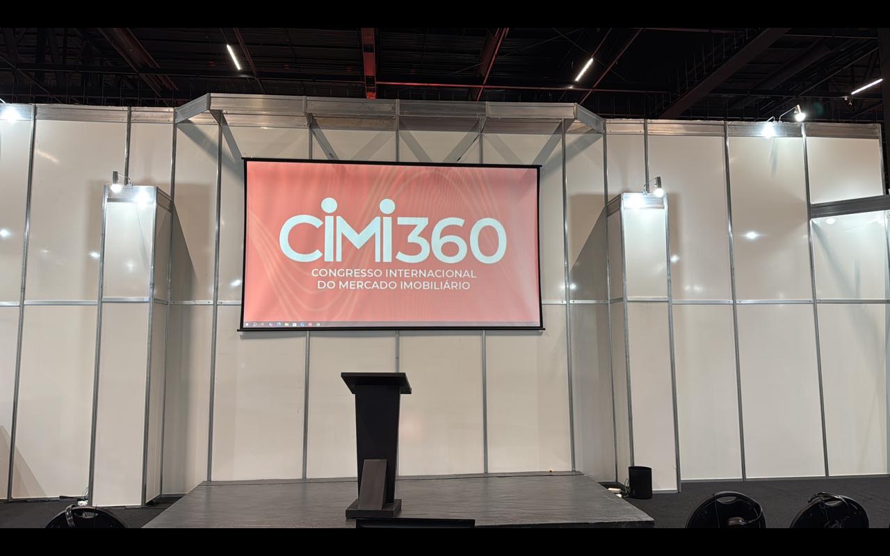 Sala modular de congresso ainda vazia, tela de projeção com a marca CiMi360, púlpito sobre praticável baixo e paredes de octanorm sob teto industrial