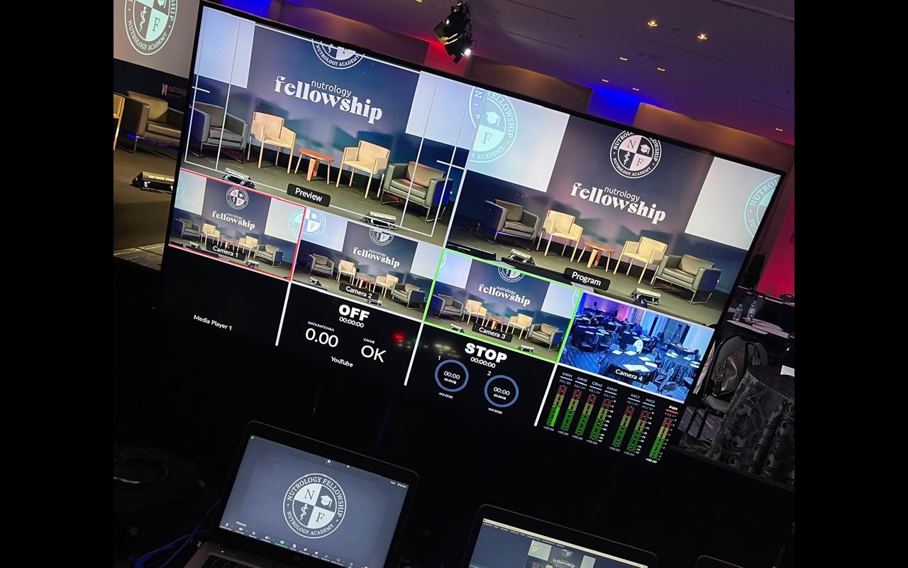 Mesa de corte no fundo do salão: um multivisor divide preview, program e quatro câmeras apontadas para o palco ainda vazio, com notebooks abaixo.