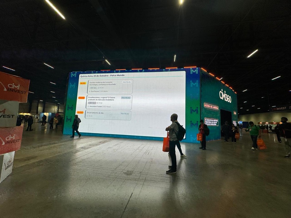 Pavilhão de eventos em penumbra, com painel de LED de grande porte exibindo a grade de programação e público circulando pelo piso livre