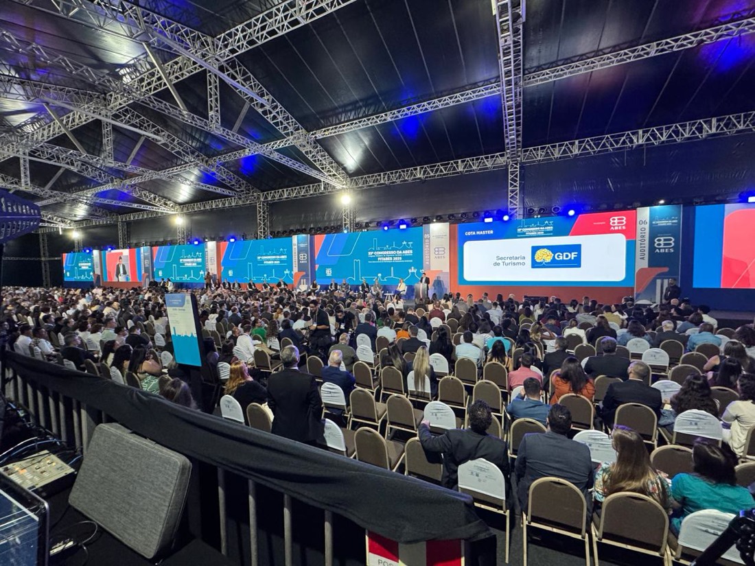 Plenária dentro da tenda do congresso, com treliça metálica aparente, painéis de LED de sinalização das salas e plateia ocupando as cadeiras